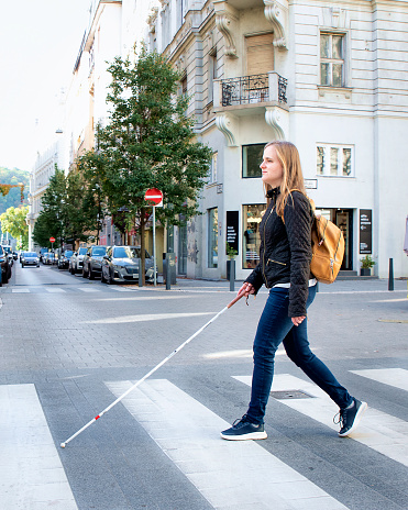 Portrait of blind woman with white cane crossing on the road in the city. A visually impaired woman wearing casual clothes and using her cane to cross the street. Full length shot.