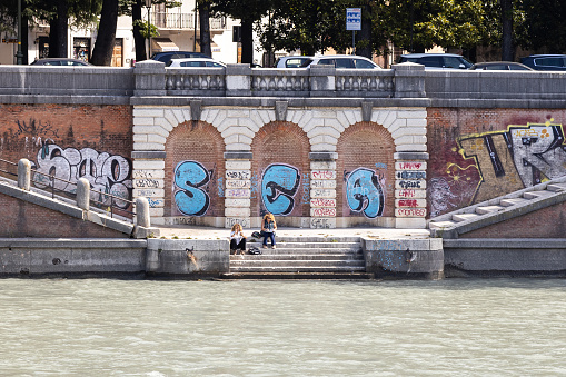 Verona, Veneto, Italy - Sep 28th, 2023: People sitting on the steps by the Adige River. Graffiti and street art on the waterfront wall