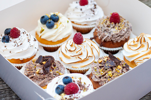 Assorted cupcakes in gift box with chocolate, vanilla and Caption:\tcream, whipped meringue, raspberry, blueberry and bites of chocolate