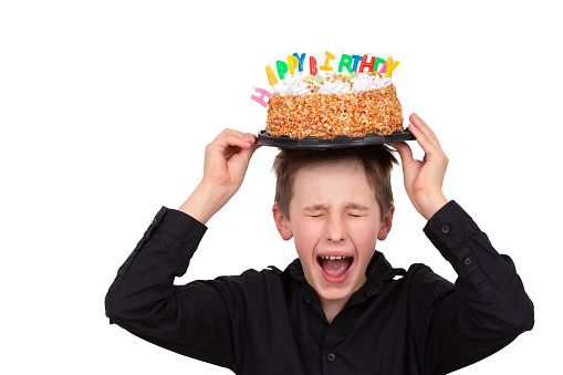 A teenage boy holds a birthday cake on his head and screams, he doesn't want to grow up. A child celebrates his birthday on a white background.
