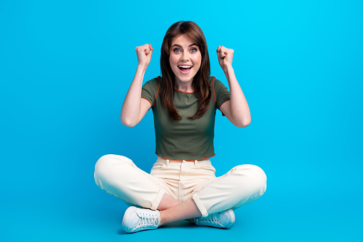 Full length photo of ecstatic girl dressed khaki t-shirt white pants sit on floor win bet scream yeah isolated on blue color background.