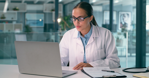 Doctor, woman and computer for healthcare research, hospital analysis and typing report or review with glasses. Medical worker or young person reading clinic charts or information on laptop in office