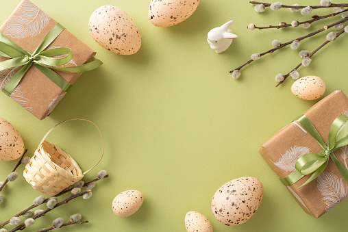 Picturesque Easter tableau idea. From top, view of paper packages, porcelain rabbit figurine, speckled eggs, basket, fresh willow boughs, on a soft green stage with empty space for script or publicity