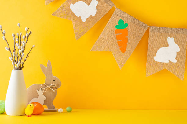 istock Easter celebration scene, side view of table set with toy rabbits, eggs in various hues, baby chick, vase holding pussy willows, rustic pennant garland, all against orange wall, with area for text 2040342122