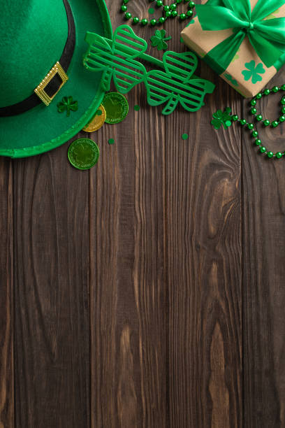 istock Charming St. Patrick's vertical top view arrangement, presenting shamrocks, fairy hat, lucky coins, surprise, fun eyewear, festive beads, confetti on a wood setting, with a spot for text or promotion 2040342636