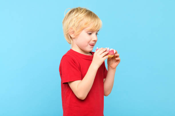 cute little boy with blond hair and freckles having fun with glazed donuts, sticking tongue out. children and sugary junk food concept. boy holding colorful donuts, eating junk unhealthy food full of sugar, isolated on pastel blue studio background. - preschooler caucasian one person part of ストックフォトと画像