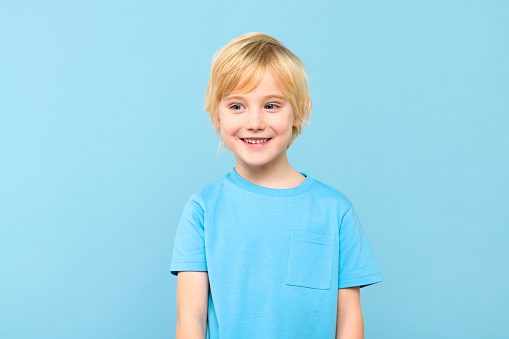 Happy young caucasian preschooler boy in casual outfit smiling, looking to the side, isolated over pastel blue background.