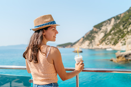 Young woman drinking iced coffee while looking at view from the boat on the Ithaca island in Greece.