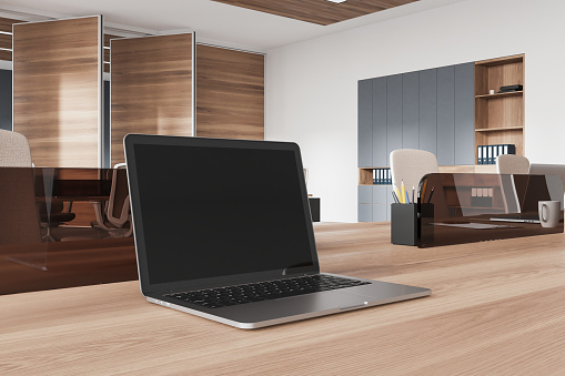 Corner view of workplace interior with black laptop screen, side view wooden table. Closeup of office work zone with technology and furniture. 3D rendering