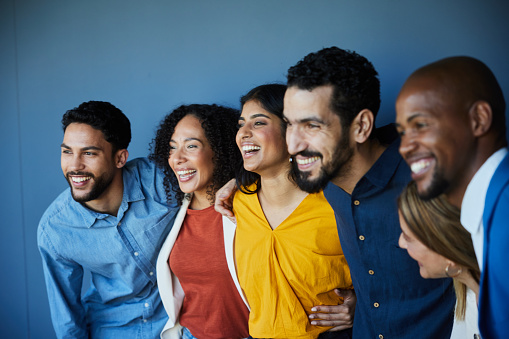 Laughing group of diverse businesspeople standing arm in arm together in front of a blue office wall