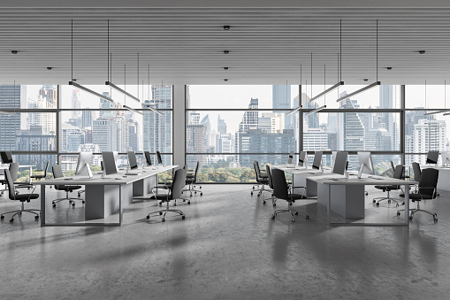 Modern coworking interior with seats and pc desktop, shared table on grey concrete floor. Office business workspace and panoramic window on Bangkok skyscrapers. 3D rendering