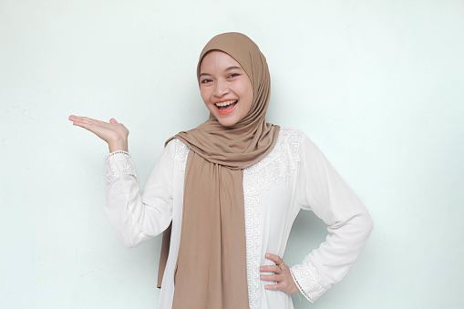 Young Asian Muslim woman wearing hijab, smiling while pointing and showing to copy space beside her. Isolated in gray background.