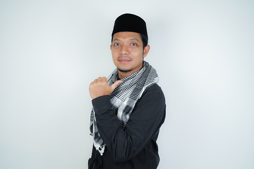 Cheerful handsome Asian Muslim man wearing Arab turban sorban pointing hand finger at empty space for advertising on isolated background
