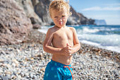 Little boy with blond hair in swimming shorts on the seashore on a summer sunny day.