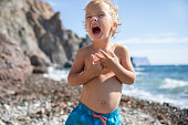 A beautiful blond child 2.5 years old in swimming trunks at the sea on a sunny warm summer day. A little boy screams loudly and plays on the rocky ocean shore.