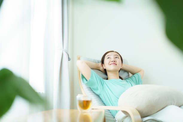 Delightful Asian middle adult woman relaxing and sitting on the cozy lounge chair and drinking a cup of tea in the morning. Beautiful - good looking Asian young woman enjoying a cup of hot tea in morning.