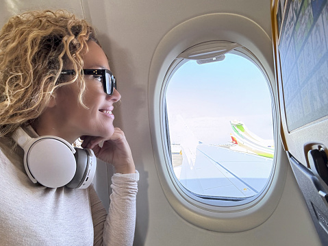 Side view of female passenger inside airplane flight looking outside the window the wing and blue sky smiling and enjoying trip listening music on headphones. Travel and holiday vacation people happy