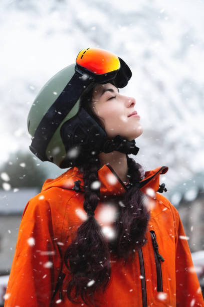 Smiling young woman, portrait, standing and enjoying snowfall, wearing protective clothing, helmet and goggles. Winter sports resort stock photo