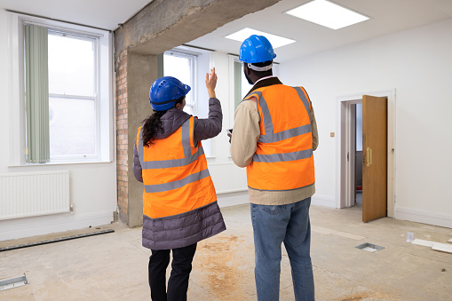Three quarter length rear view of a mid adult male and young female construction workers discussing building plans together as a team for the project they are working on in the room they are planning on renovating. They're wearing casual clothing and have their hi vis jackets on as well as hard hats.