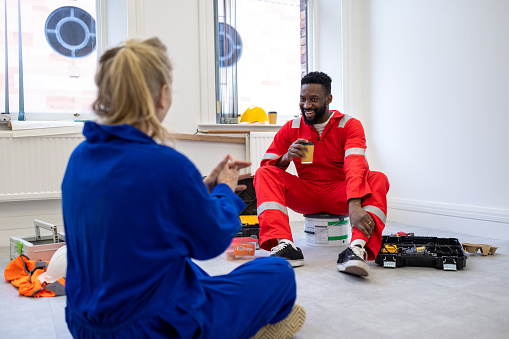 Over the shoulder view of a mid adult male and mature female building contractors sitting down on paint cans taking a break in a room they are renovating, they are enjoying their lunch and hot drinks. They're wearing reflective coveralls and having a chat on their lunch break.