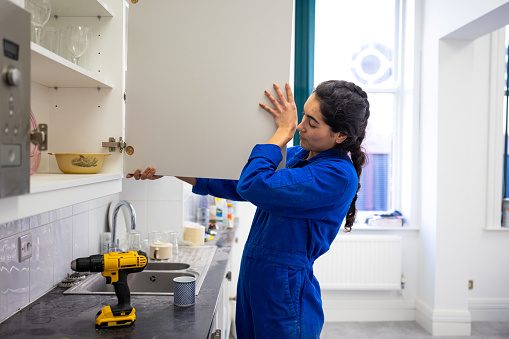 Three quarter length side view of a young female blue collar worker wearing coveralls, she is attaching a new cabinet door in an office kitchen in Newcastle, England. There is a drill on the kitchen counter.