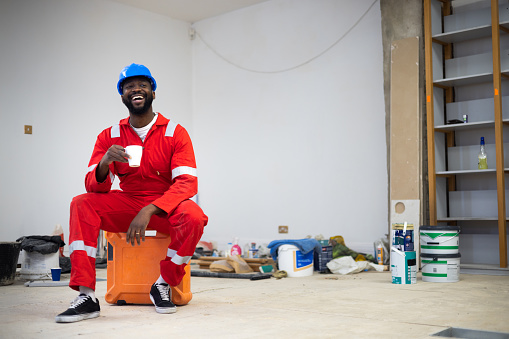 Full length wide view of a mid adult male building contractor sitting down on a toolbox taking a break in an office he is renovating, he is enjoying a hot drink. He's wearing a hard hat and reflective coveralls.