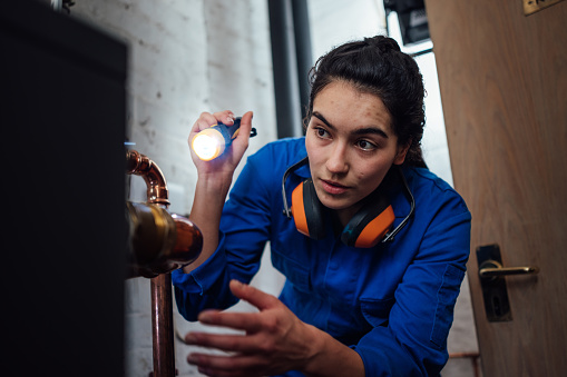 Front view waist up of a young female gas engineer/technician wearing coveralls repairing a boiler. She is holding a torch to shine a light on the pipe she is going to repair.