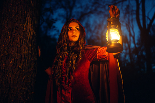 Witch in a red robe holding a lantern in the forest at night.