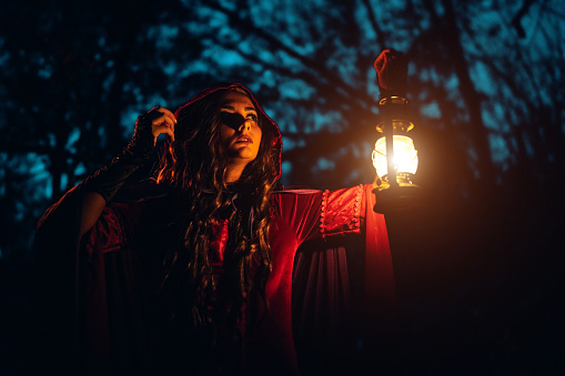 Witch in a red robe holding a lantern in the forest at night.
