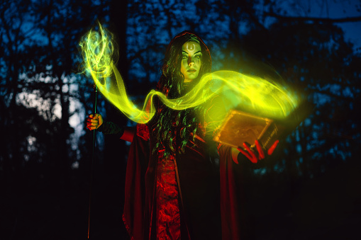 Young woman dressed like an evil witch using magic cane and a book and practicing magic in the forest at night.