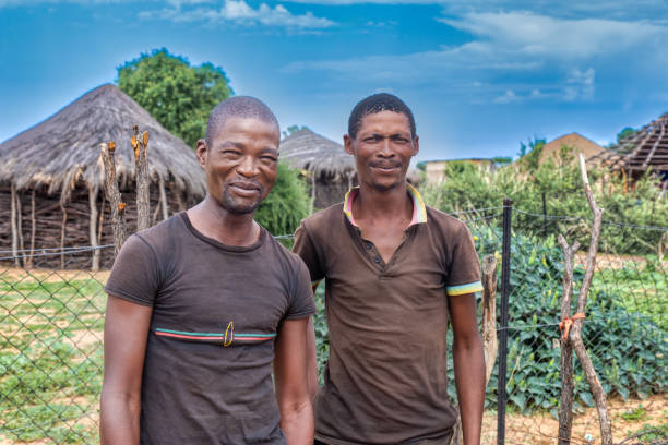 dos jóvenes y felices africanos de la aldea de pie frente a una choza con techo de paja. - bechuana fotografías e imágenes de stock