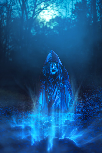 Young woman dressed like an evil witch and practicing magic in the forest at night.