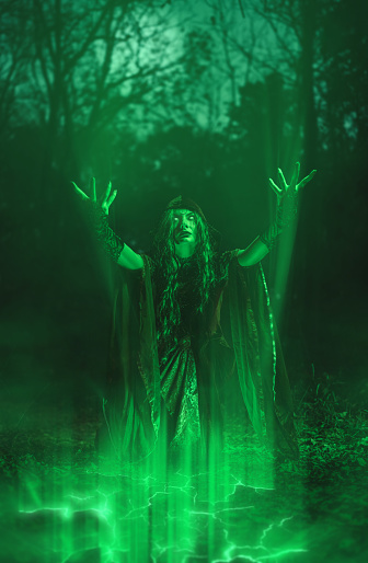 Young woman dressed like an evil witch and practicing magic in the forest at night.