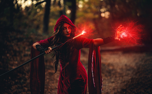Young woman dressed like an evil witch using magic cane and practicing magic in the forest at night.