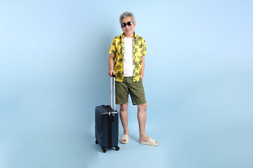 HAPPY SONGKRAN DAY. Asian tourist senior man in summer clothing and sunglasses with gesture of holding suitcase isolated on blue background. Songkran festival. Thai New Year's Day.