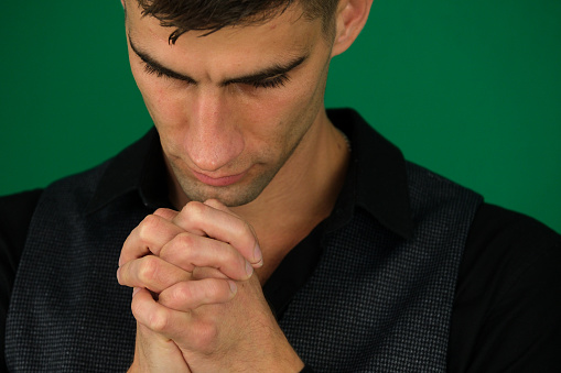 A young man with folded hands praying in closeup. emotions of a handsome man guy on a green background chromakey close-up dark hair young man