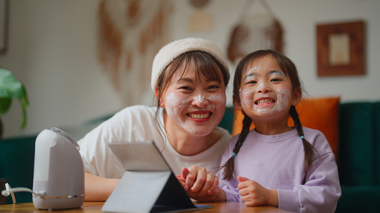 A portrait of mother is enjoying doing beauty skin care and relaxing with her small daughter in the living room at home.