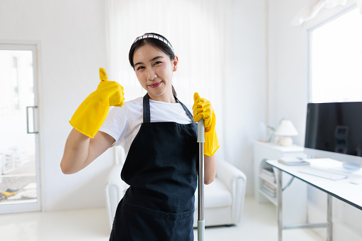 Portrait of a young Asian woman cleaning the house, standing holding a mop and smiling happily.