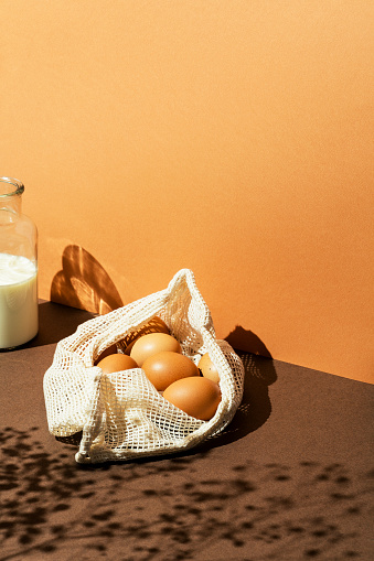 Eggs in a mesh bag with milk in glass bottle on brown background. Sun light effect with floral shadows, high angle shot. Copy space background.