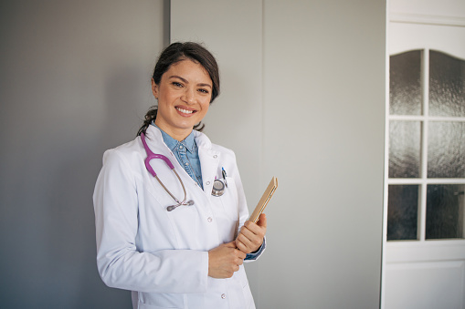 Portrait of female doctor standing in medical cabinet. She is smiling at the camera. Wearing a stethoscope around her neck and holding a notebook in her hands. Healthcare and medicine. Front view.