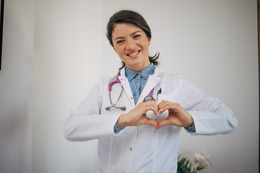 Portrait of beautiful female doctor in white uniform showing heart with hands. She is smiling and looking at the camera.