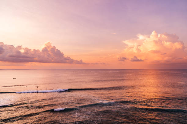 A captivating aerial view captures the warm colors and textures of nature during sunset. The ocean’s gentle waves crash into a barrier in the foreground, while the sky is painted with hues of orange and purple. Soft clouds scatter across the horizon. Shot taken on Bali.