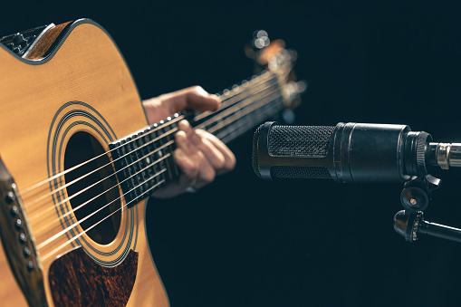 Male musician playing acoustic guitar behind microphone in recording studio. The concept of music recording, rehearsal or live performance.