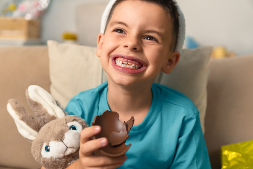 Fun brazilian boy with big smile eating easter chocolate egg at home, in living room