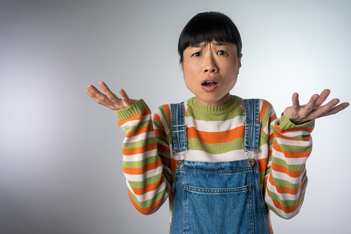 Portrait of a confused Asian casual girl shrugging shoulders isolated over grey background. Confused Japanese woman shrugs her shoulders in a clueless gesture.