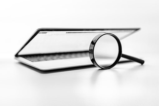 Laptop computer with magnifying glass as a symbol for searching information on the internet.Black and white image on a white background with selective focus on the glass of a magnifying glass that is upright on the laptop