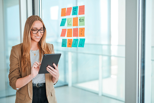 Businesswoman wearing eyeglasses and brown jacket, smiling happily, working and swiping digital tablet, standing beside glass wall with sticky notes in modern office