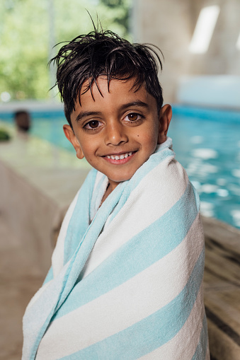 Close-up of a young boy wrapped in a light blue and white swimming towel. His hair is wet from being in a swimming pool. He is looking straight into the camera whilst smiling.