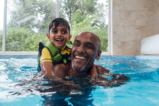 Close-up of grandad and grandson playfully swimming together in a swimming pool. They are both smiling as the young boy is perched on his back as the man is swimming. The boy is wearing a green life jacket.