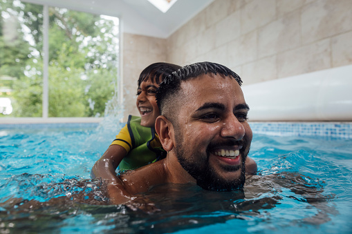 Close-up of father and son playfully swimming together in a swimming pool. The son is riding on the father's back whilst laughing. The man is smiling as he swims along.  The child is wearing a green life jacket.